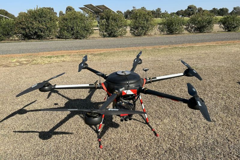 Six rotor agricultural drone sitting on gravel beside a paddock and line of trees
